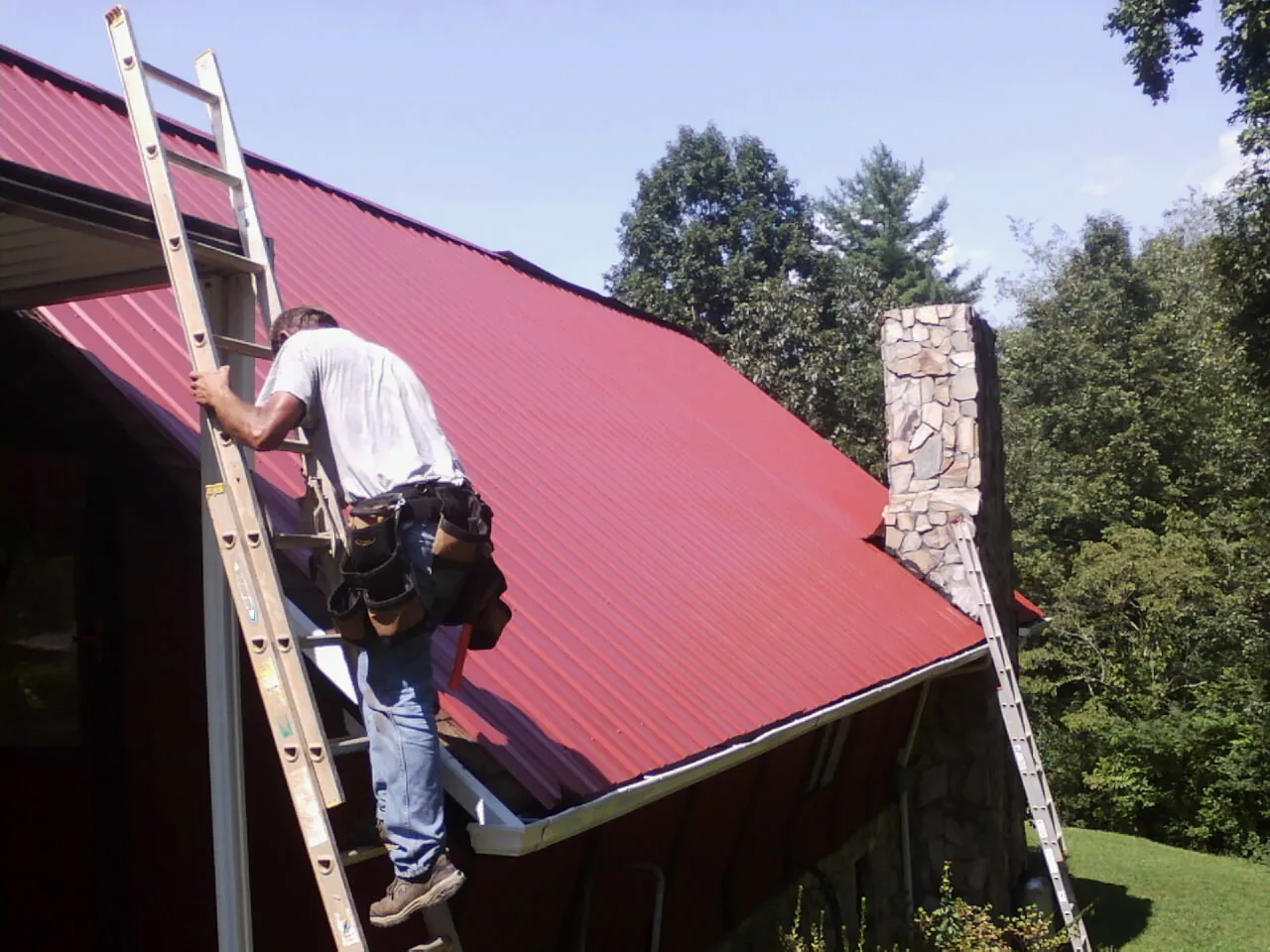 Roofer on a ladder beside a red metal roof and stone chimney — Soffit & Fascia Repair by Roofing Brothers in Hillsboro
